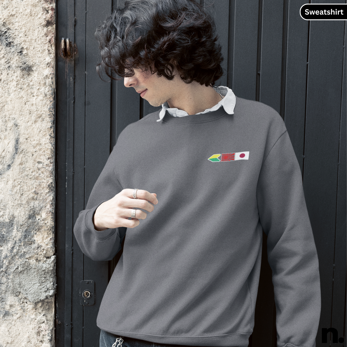 Person wearing a gray sweatshirt with a logo, standing against a dark door.