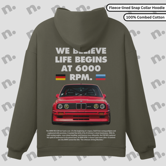 Flat lay of a premium snap-collar hoodie featuring a BMW E30 M3 design and the text "Life Begins at 6000 RPM", paired with minimalist accessories on a clean background.