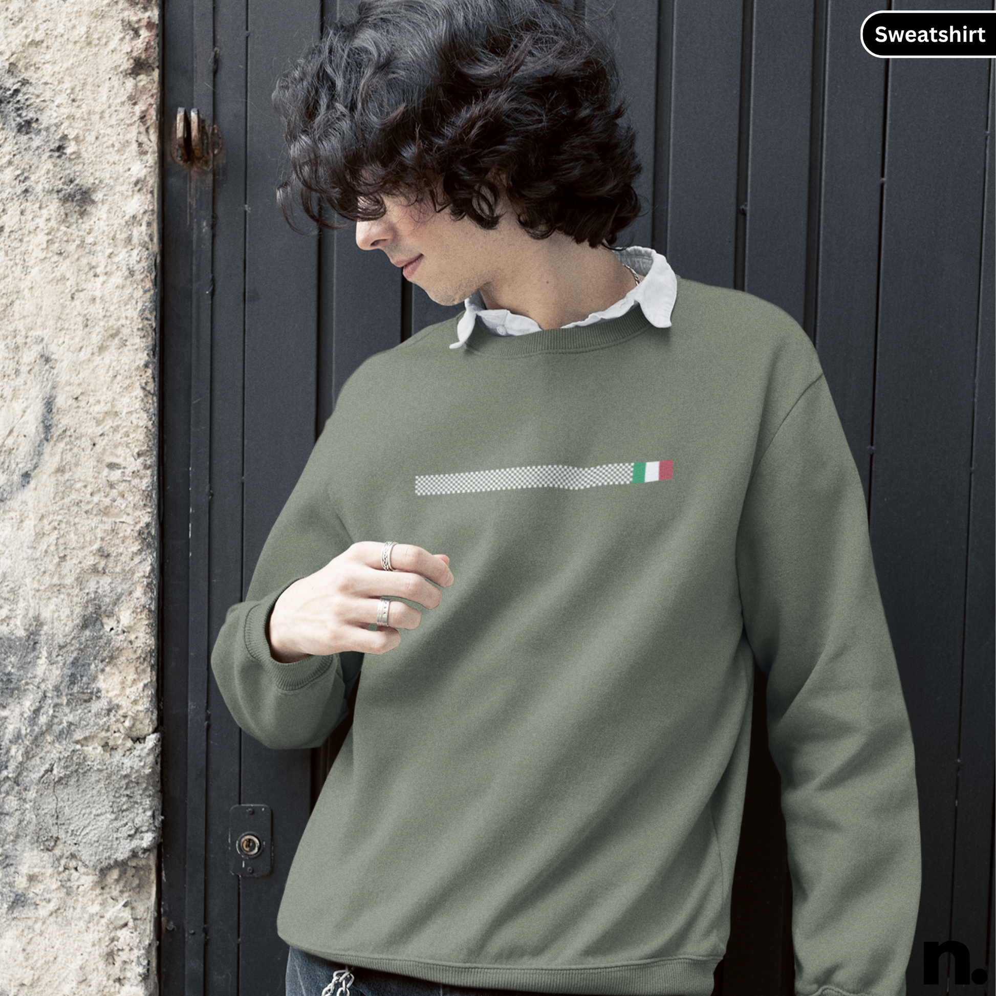 Person wearing a green sweatshirt with a small flag design, standing against a dark door.