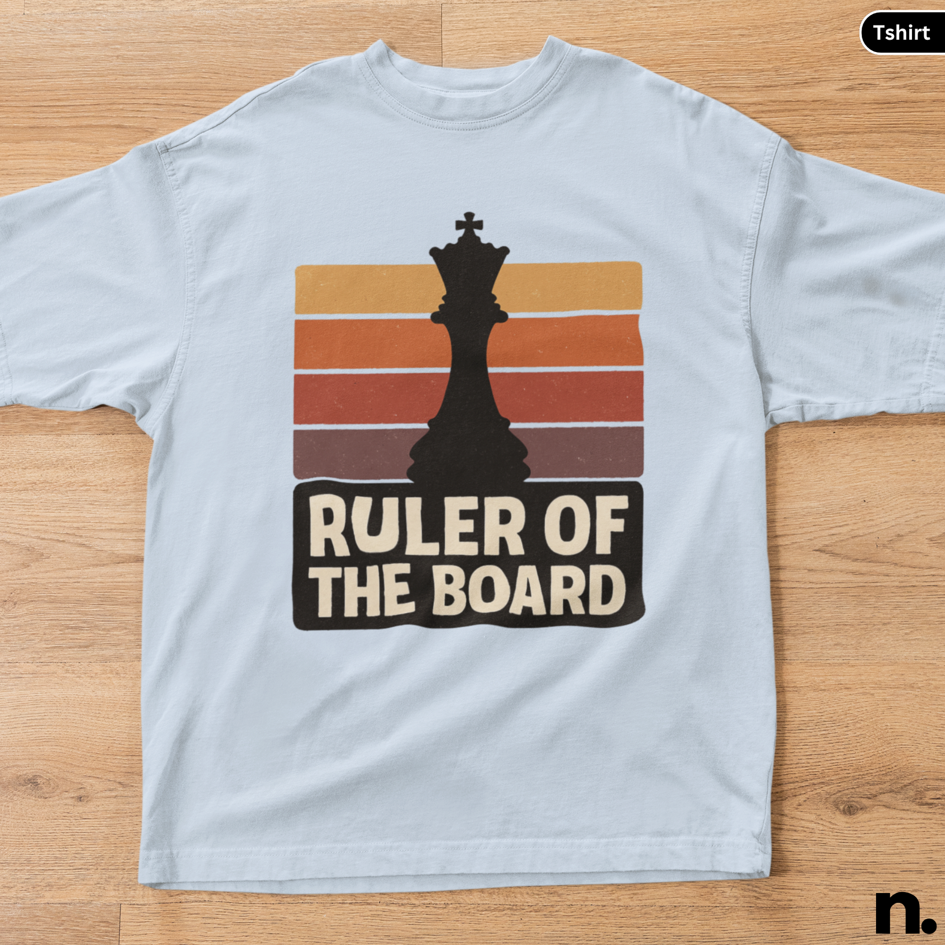 White t-shirt with a chess-themed graphic and text 'Ruler of the Board' on a wooden surface.