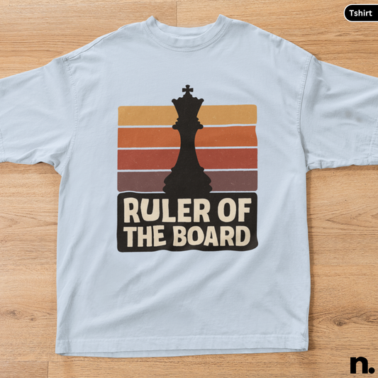 White t-shirt with a chess-themed graphic and text 'Ruler of the Board' on a wooden surface.