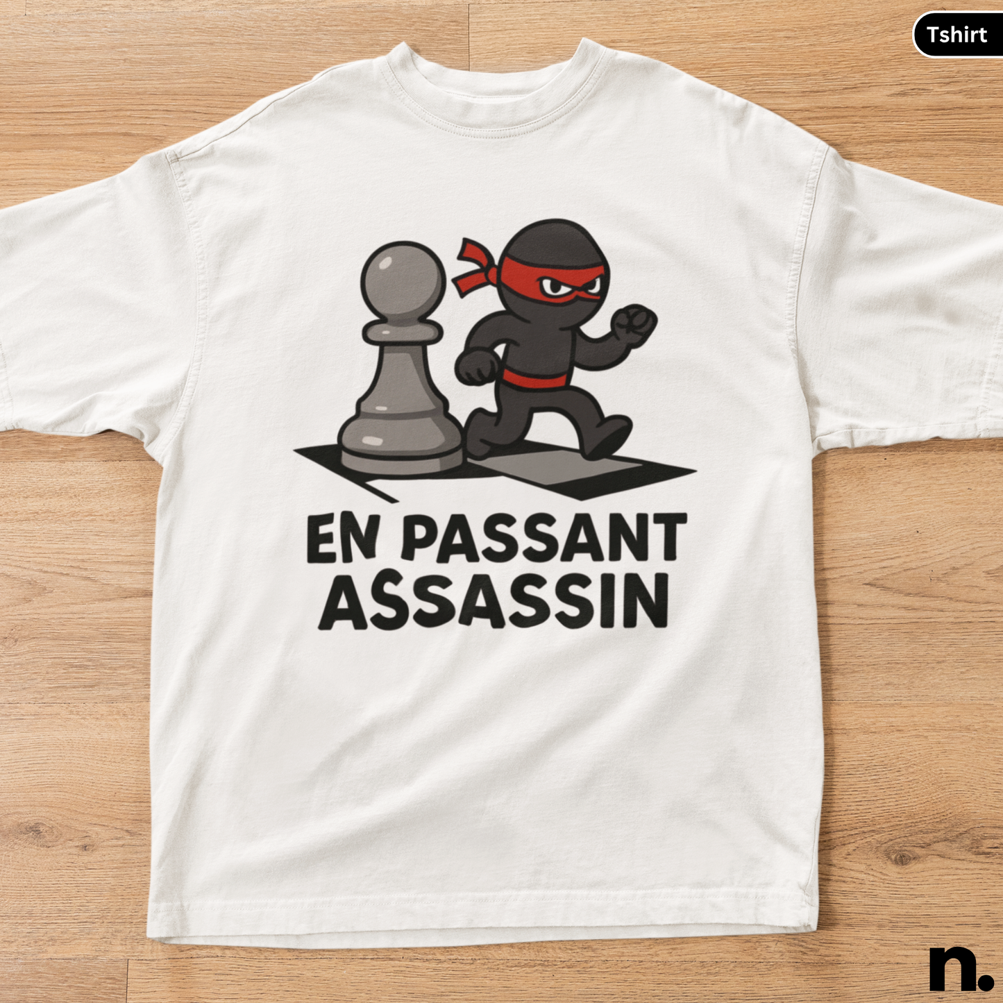 White t-shirt with a graphic design of a ninja and chess piece, text 'EN PASSANT ASSASSIN', on a wooden surface.