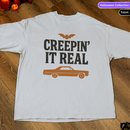 White t-shirt with 'Creepin' It Real' text and car graphic on a wooden surface, labeled as Halloween Collection.