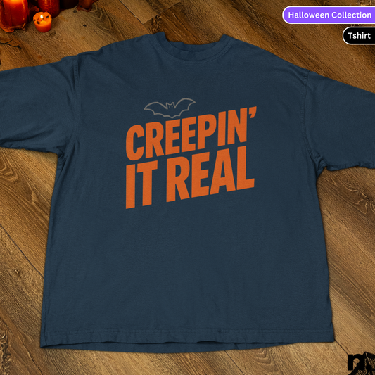 Navy blue t-shirt with orange text 'CREEPIN' IT REAL' on a wooden surface, part of a Halloween Collection.