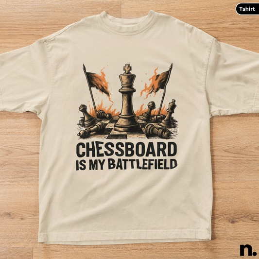 Beige t-shirt with a chess-themed graphic and text 'Chessboard is my battlefield' on a wooden surface.